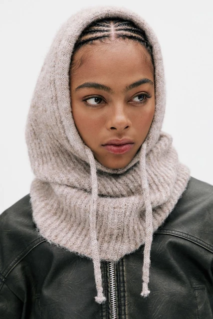 A woman poses indoors wearing a knitted balaclava with drawstrings and a black leather jacket against a white background in a studio shot.