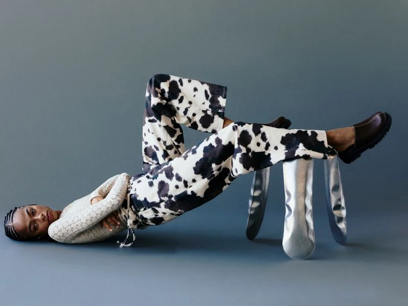 Woman in cow-print pants rests legs on silver stool against grey backdrop. She wears a cream sweater and dark brown shoes in studio shot.