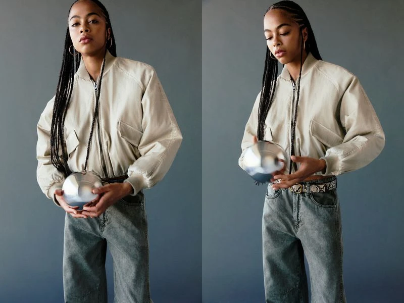 A young woman with braided hair holds a silver sphere-shaped handbag, wearing a beige jacket and grey jeans, in a studio setting.