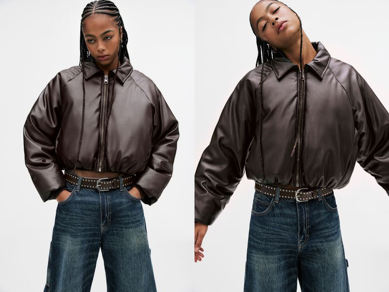 Eye-level shot of a woman with braided hair in a brown cropped jacket, jeans, and studded belt, posing in a photo studio.