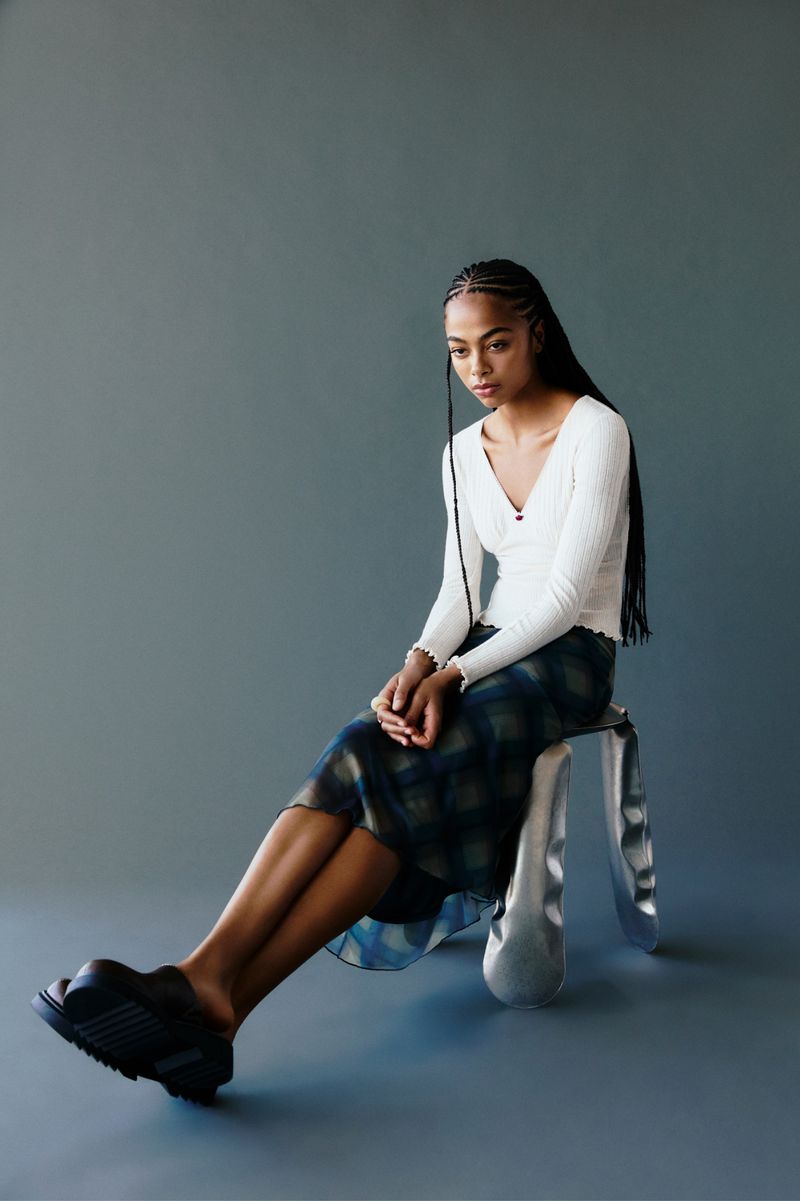 A young adult female sits on a silver stool in a studio, wearing a white top, plaid skirt, and platform shoes against a gray background.