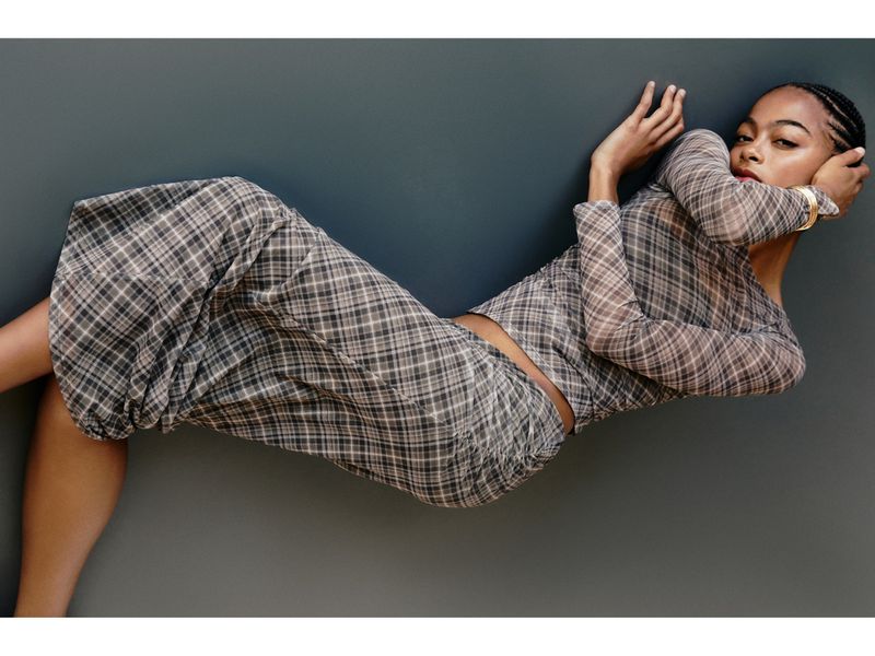 Young woman in grey plaid top and skirt set posing against grey studio wall. Bracelet accessory on wrist, red lipstick, braids visible.