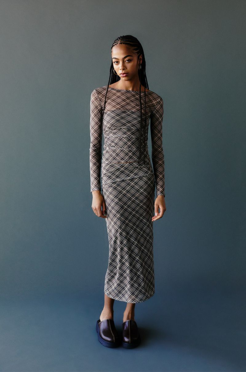 A slender African American woman stands indoors wearing a plaid dress with long sleeves and black shoes against a solid grey background.