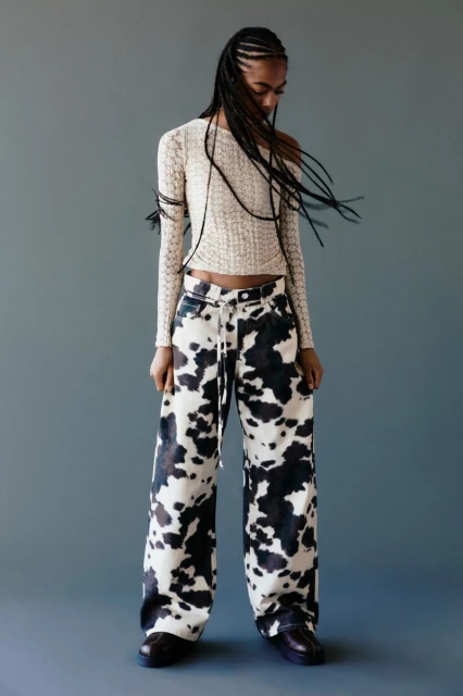 A young woman with braided hair poses in a photo studio wearing a knitted top and cow-print jeans against a gray background.