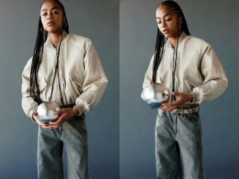A woman wearing a beige bomber jacket and grey jeans holds a shiny silver sphere against a solid grey studio background.