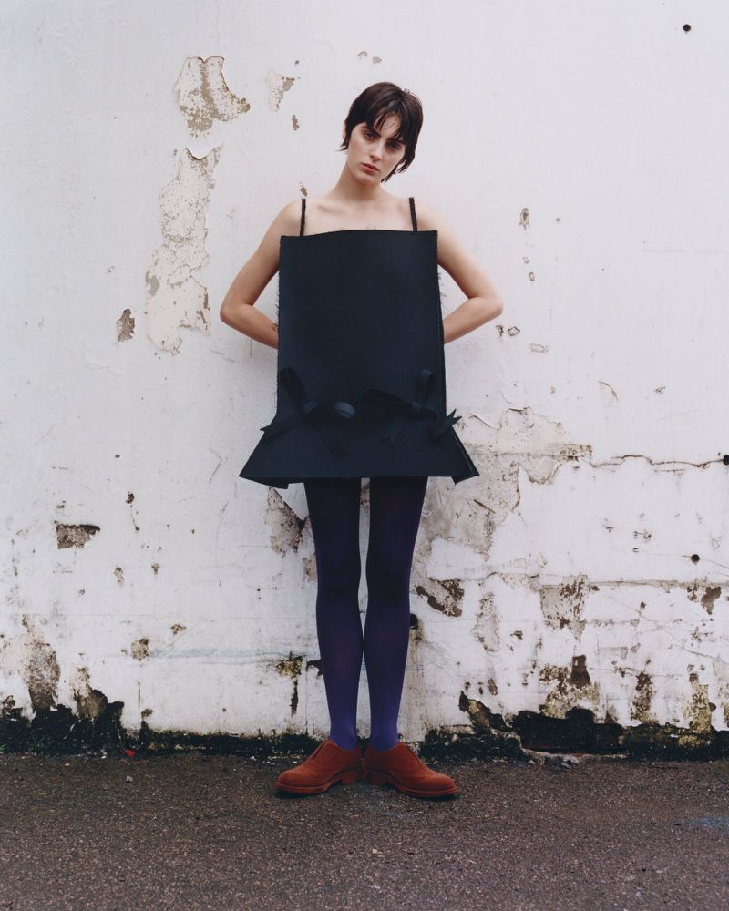 Fashion shot of a young female adult standing with hands on hips, wearing a black square dress and purple tights against a grunge wall.