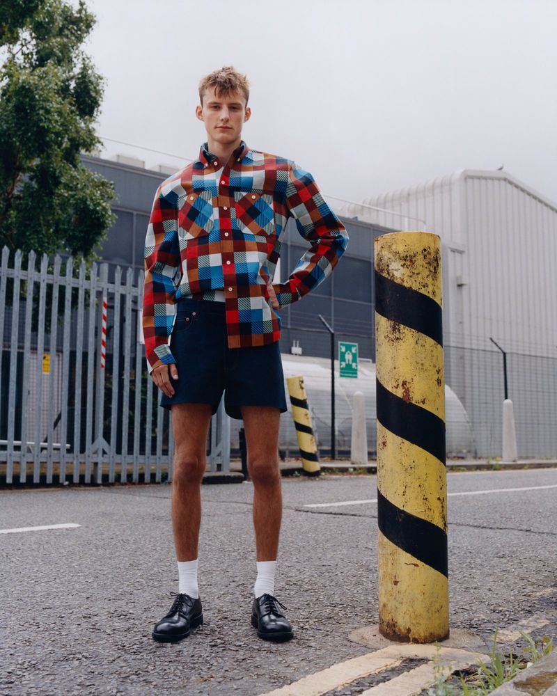 Young man poses in a multicolor plaid shirt and navy shorts on a city street, near a gray fence and striped yellow bollard.