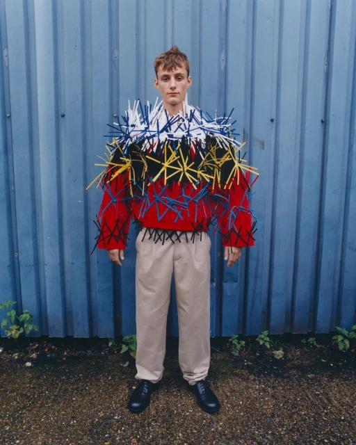 Male fashion model poses outdoors against a blue metal wall wearing a red and black sweater with multi-colored straw-like decoration.