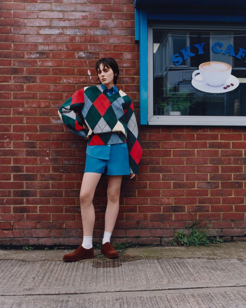 A young adult female poses against a brick wall in an argyle sweater and blue shorts, showcasing an editorial fashion shot outdoors.