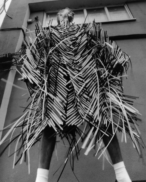 Black and white portrait of a person wearing a unique straw-like garment in an outdoor urban setting, low angle, and interesting texture.