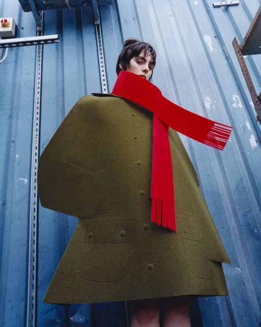 Stylish adult female posing in an olive green coat with a bright red scarf in front of a corrugated metal industrial wall outdoors.