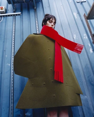Model wearing an olive green sculptural coat and bright red scarf against a blue corrugated metal wall.