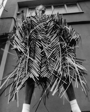 Model wearing a structured garment made of fringe strips standing against a building exterior.