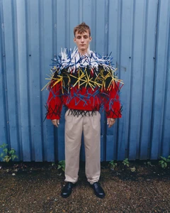 A man wearing an avant-garde red sweater embellished with protruding sticks standing before a blue corrugated metal wall.