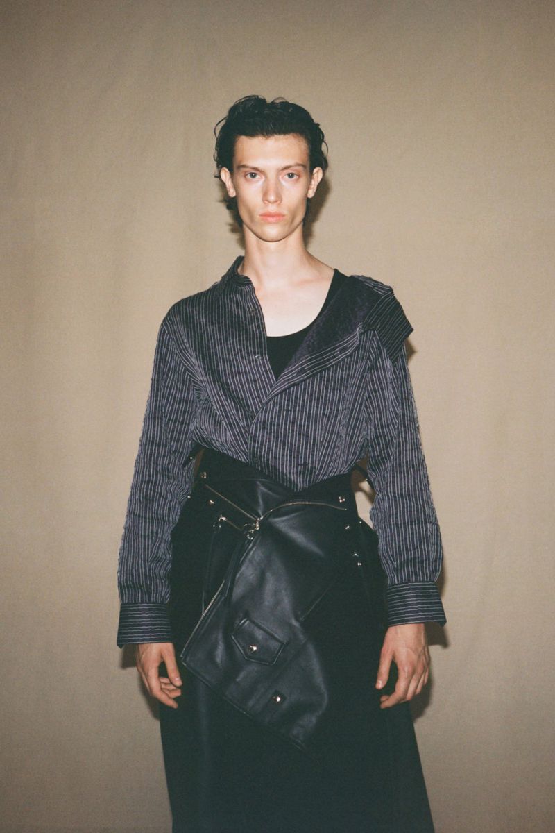 Stylish male model posing in a pinstripe shirt and avant-garde leather skirt with pockets against a neutral backdrop in a studio setting.