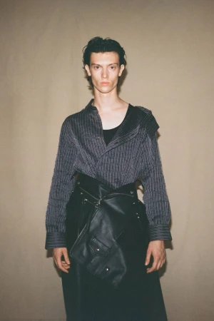 Stylish male model posing in a pinstripe shirt and avant-garde leather skirt with pockets against a neutral backdrop in a studio setting.
