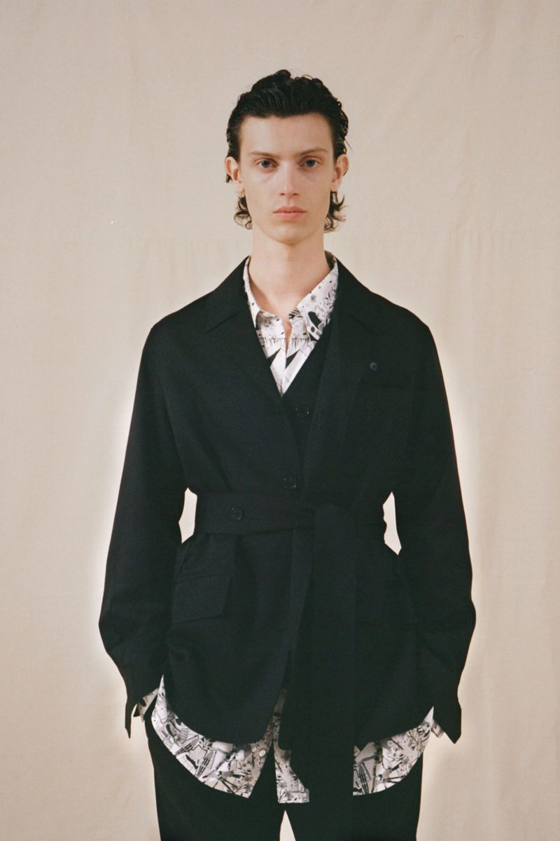 Full-length shot of a model in a black jacket with a belt, layered over a patterned shirt, standing in a neutral indoor setting.