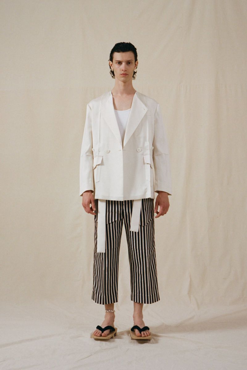 Full shot of a young adult man in a white jacket, top, and striped pants, posing indoors against a light background in a fashion shot.