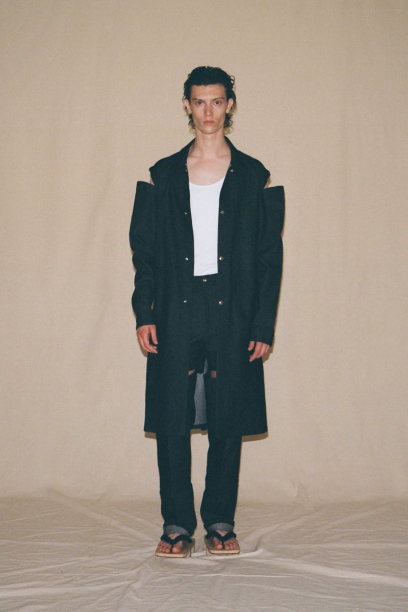 Male model in a black coat, white tank top, and black pants standing in front of a beige background in a studio environment.
