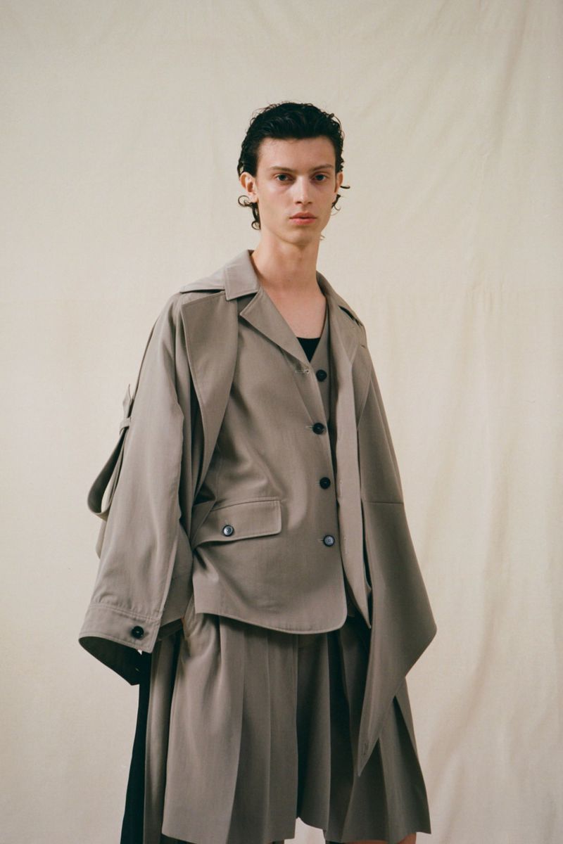 Fashionable person in an oversized grey jacket, a black top and a matching pleated skirt posing in front of a neutral background.