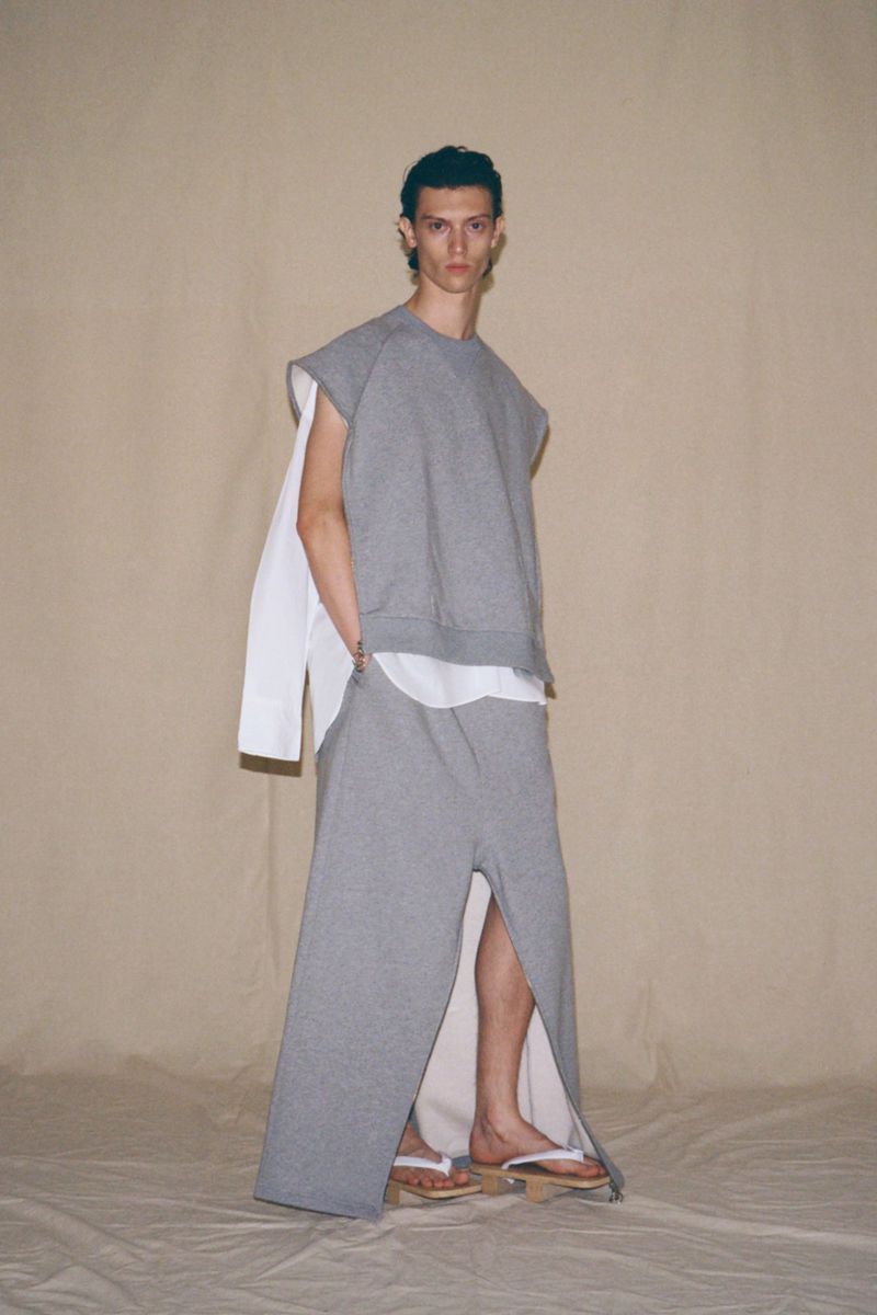 Male fashion model poses in grey sleeveless top and long skirt with white shirt layered underneath against neutral backdrop in studio.
