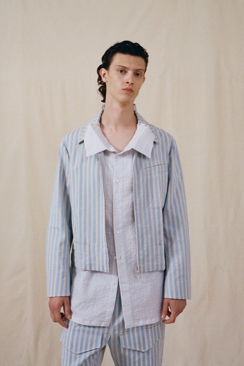 An adult male poses indoors wearing a light blue and white striped jacket, shirt, and pants against a neutral backdrop.