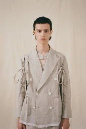 A male model wearing a light beige double-breasted blazer with side tie details against a neutral fabric background.