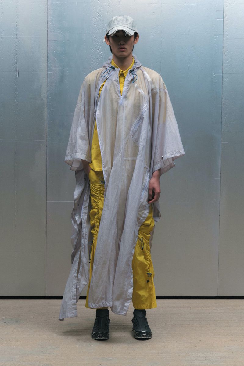 Young man in translucent poncho and cap stands against a light metallic background, showcasing fashion apparel with a modern edge.