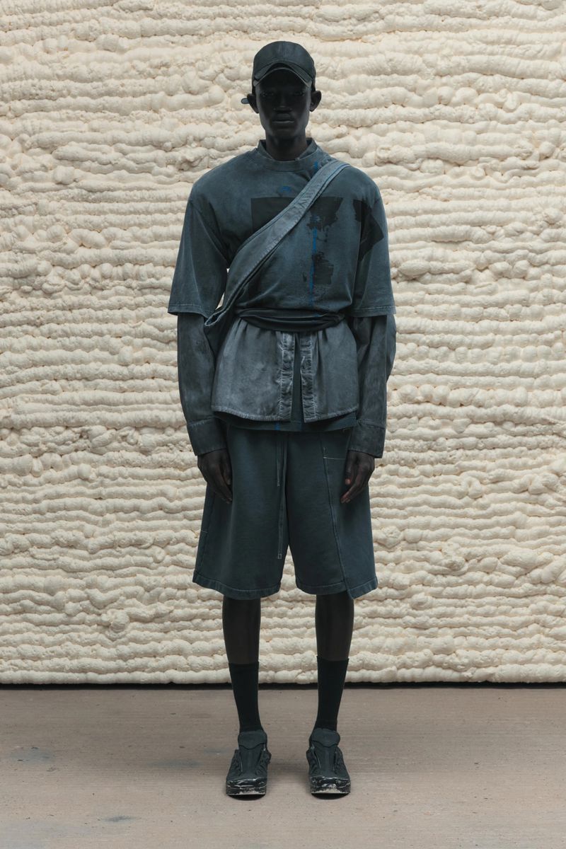 Dark-skinned man in a matching grey-blue casual outfit poses in front of a textured wall in a fashion or editorial shot.