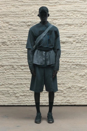 Dark-skinned man in a matching grey-blue casual outfit poses in front of a textured wall in a fashion or editorial shot.