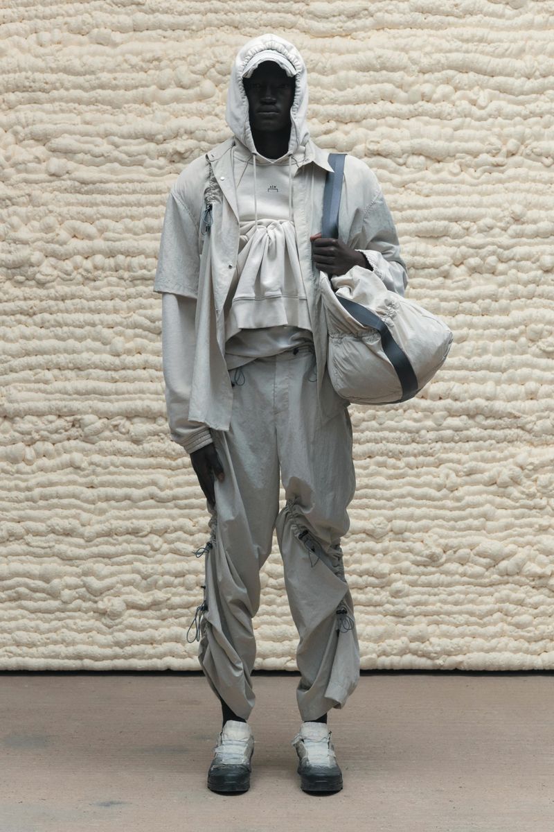 Full-length studio shot of a hooded, slender African male model in a layered tonal outfit holding a duffel bag against a textured backdrop.