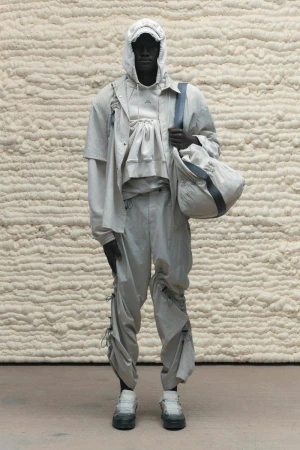 Full-length studio shot of a hooded, slender African male model in a layered tonal outfit holding a duffel bag against a textured backdrop.