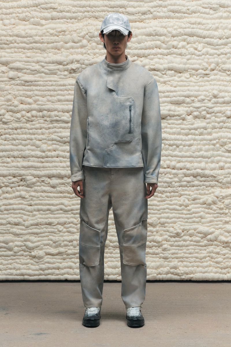 Full-length studio shot of a person posing in matching light-blue textured outerwear, pants, and cap against a matching textured background.