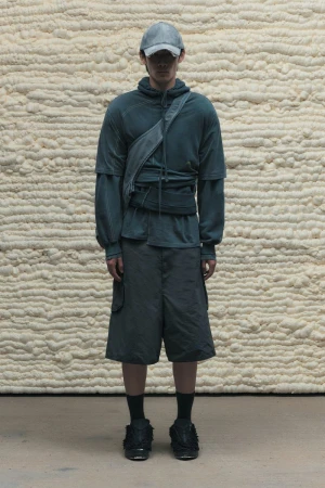 A full shot of an adult model wearing a blue-green layered outfit with a cap and crossbody bag, standing against a textured beige wall backdrop.