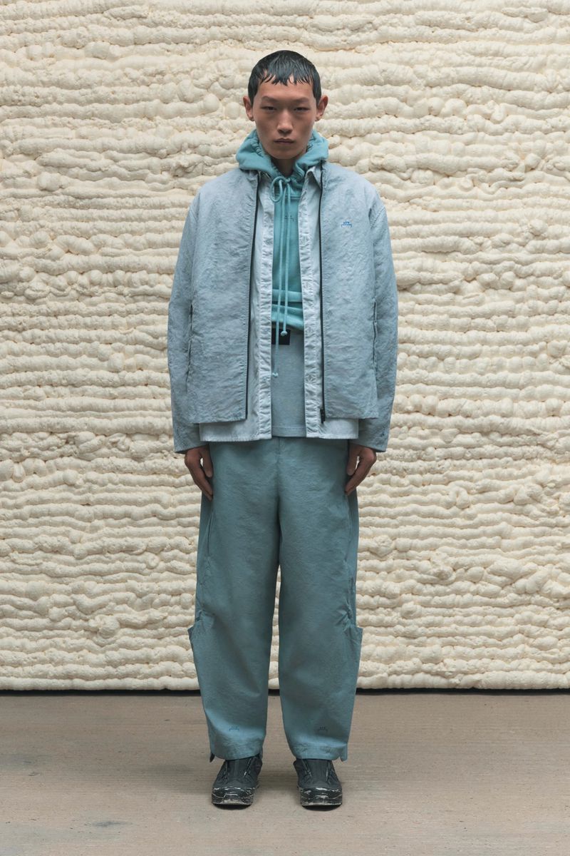 Full-length portrait of a young adult male fashion model in a light blue jacket and pants, standing indoors against a textured off-white wall.