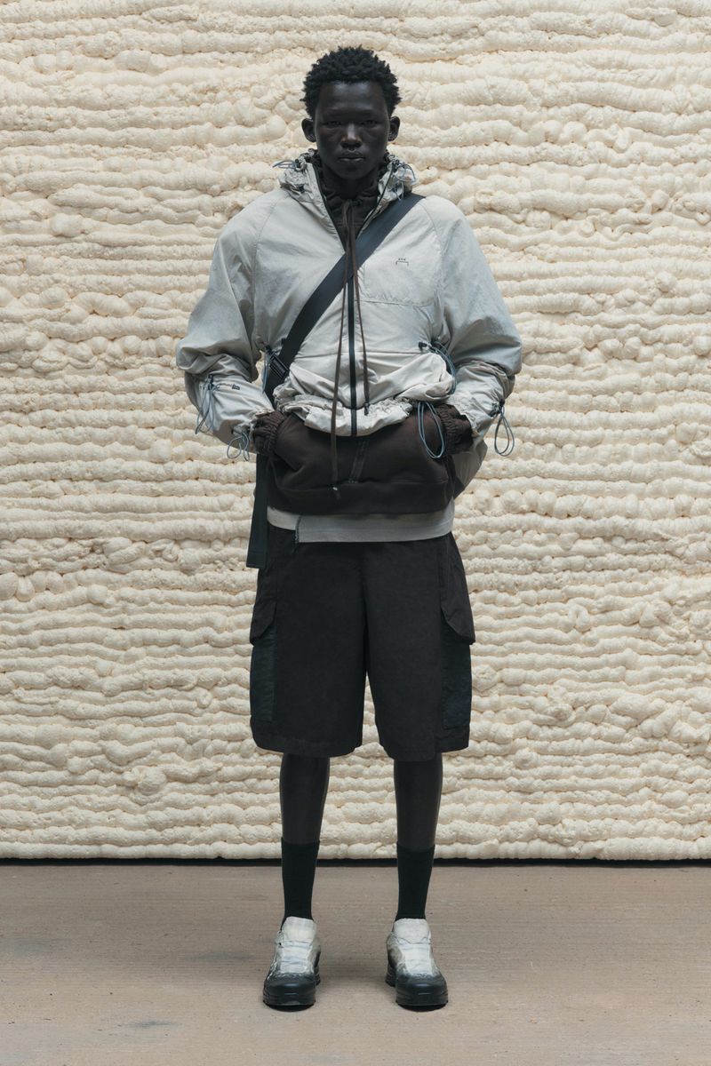Man in light jacket and dark shorts stands in front of textured backdrop in fashion shot. He is carrying a soft dark bag.