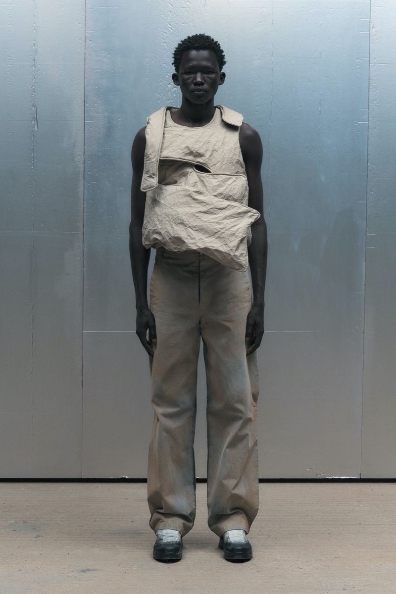 Full-length shot of a dark-skinned male model in a neutral-toned vest and pants against a grey backdrop, showcasing contemporary fashion.