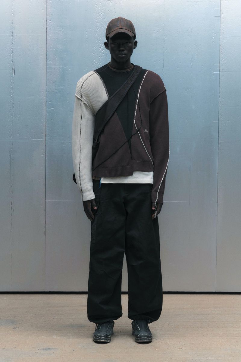 Full-length shot of a male model wearing a brown baseball cap, patchwork sweater, black pants and bag in a studio setting.