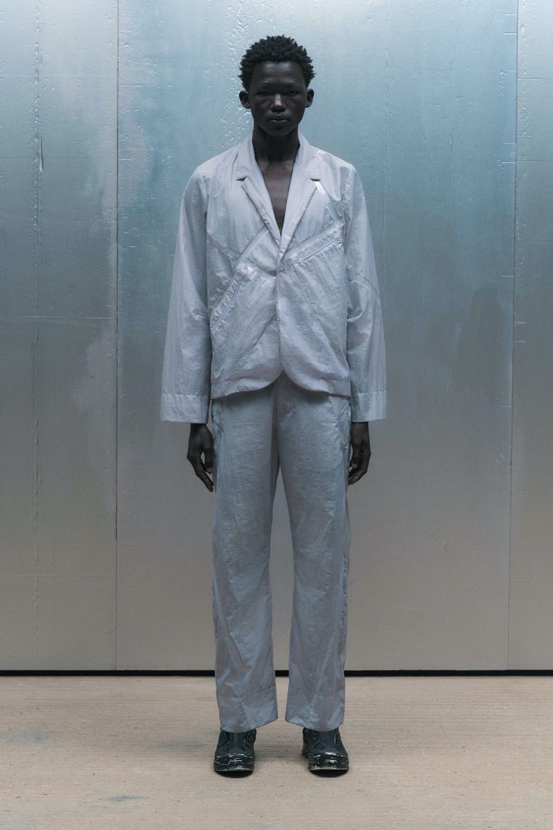 A full-length shot of an adult male model in a light grey blazer and matching pants, posing in front of a pale grey background.