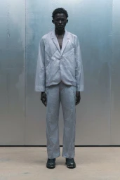 Model in a pale grey technical blazer and matching trousers standing against a metallic panel wall for A-Cold-Wall.