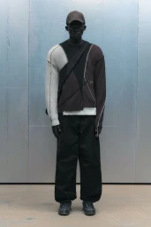 Model wearing panelled colorblock sweater, dark trousers, and baseball cap standing against industrial metal wall
