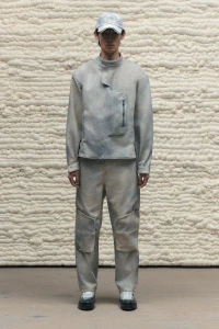 Model wearing a grey distressed jacket, matching trousers, and cap against a textured foam wall