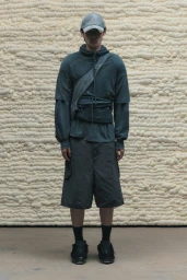 Model wearing a layered slate green hoodie, dark shorts, cap, and sneakers in front of an off-white textured foam wall.