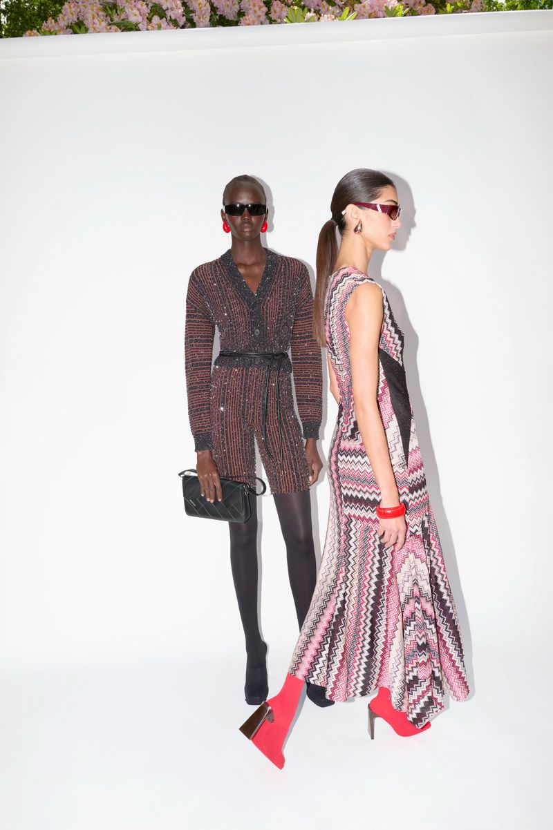 Two fashion models pose in patterned outfits against a white backdrop with flowers. One wears a cardigan set, the other a dress.