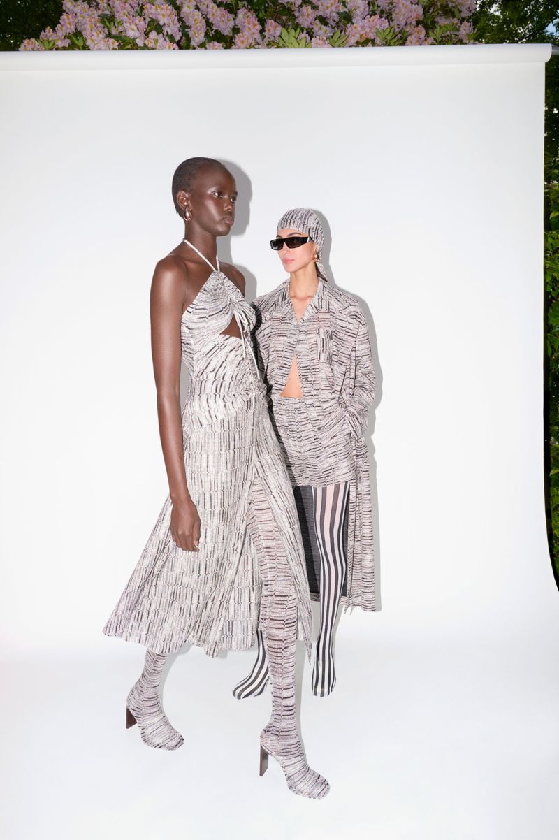 Two fashion models showcasing patterned outfits in a studio with soft, natural lighting and floral accents. Full shot with white backdrop.