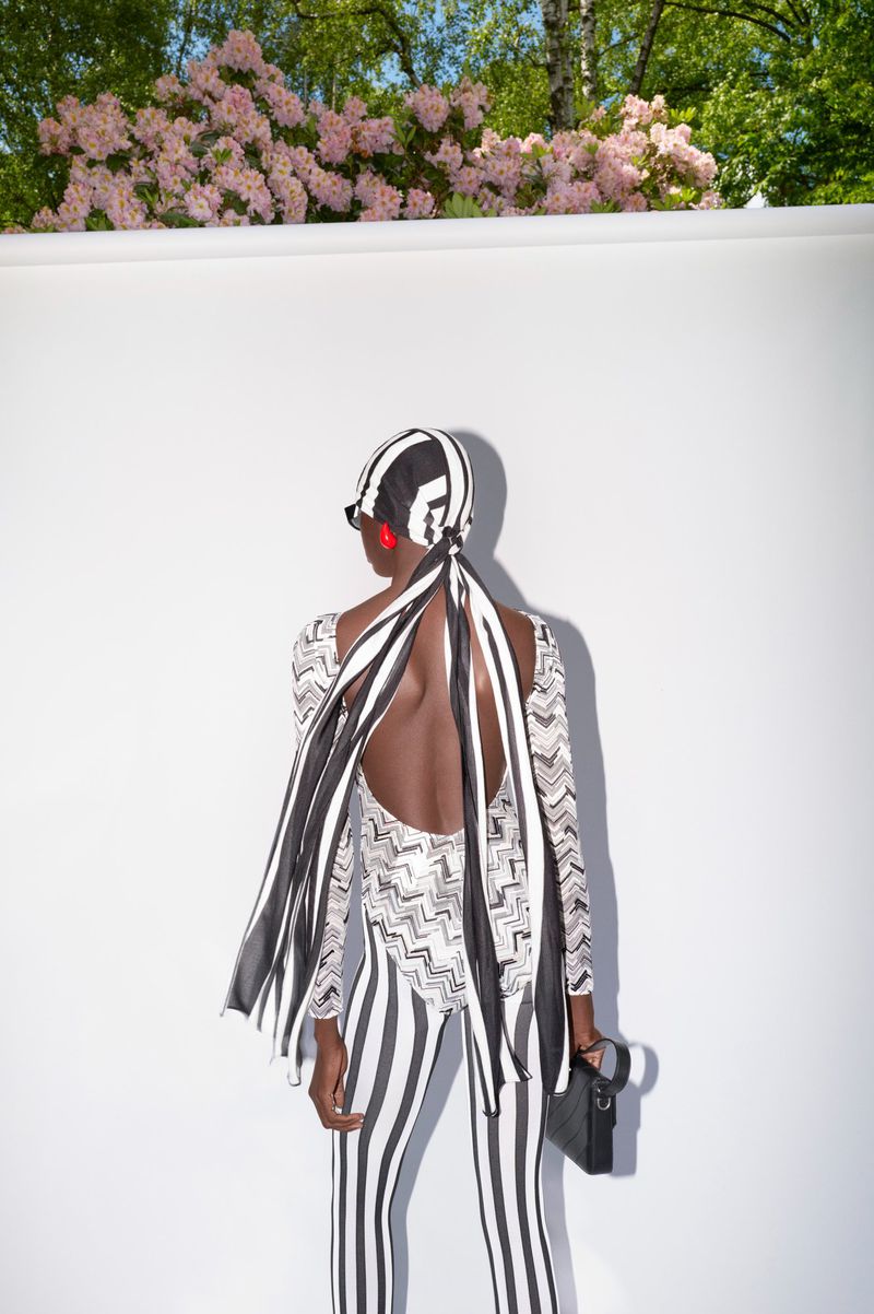Back view of a woman in a striped catsuit with a matching headscarf and purse, posing against a white wall and trees in the background.