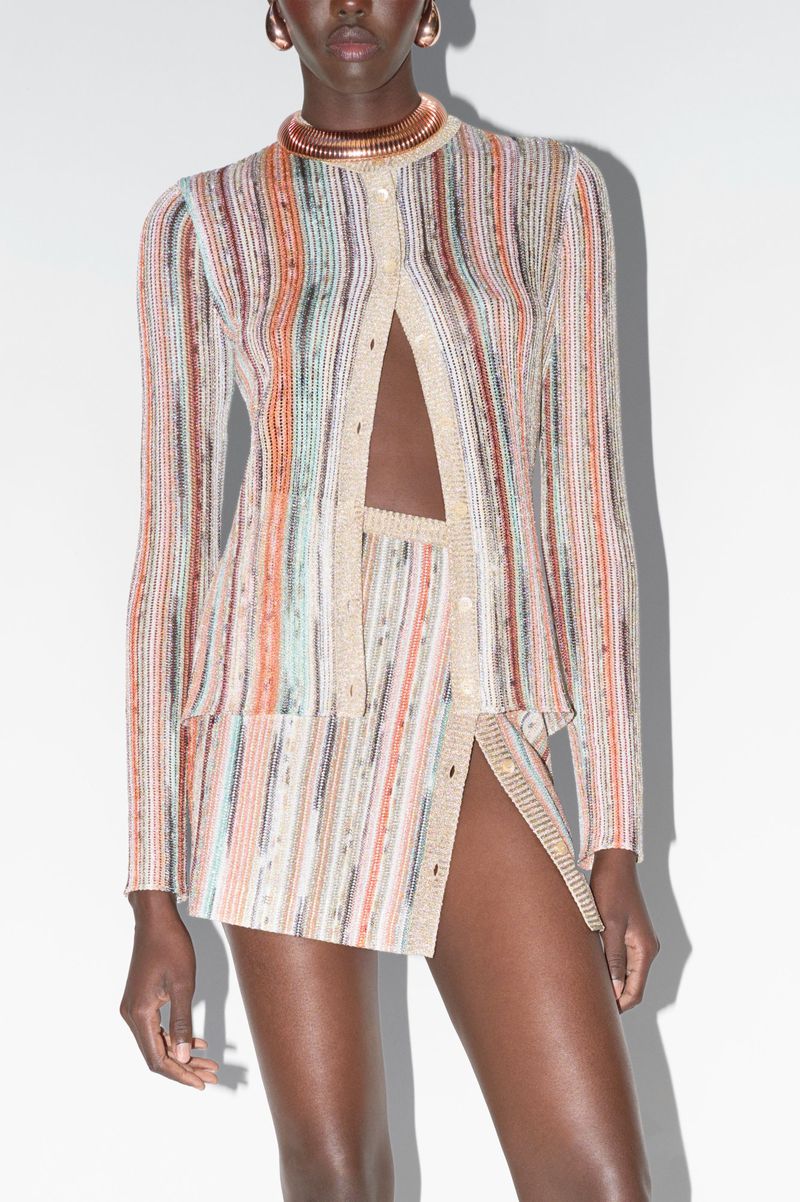 Fashion shot of a Black female model in a patterned knit skirt suit with gold collar necklace, posing in front of a white backdrop.