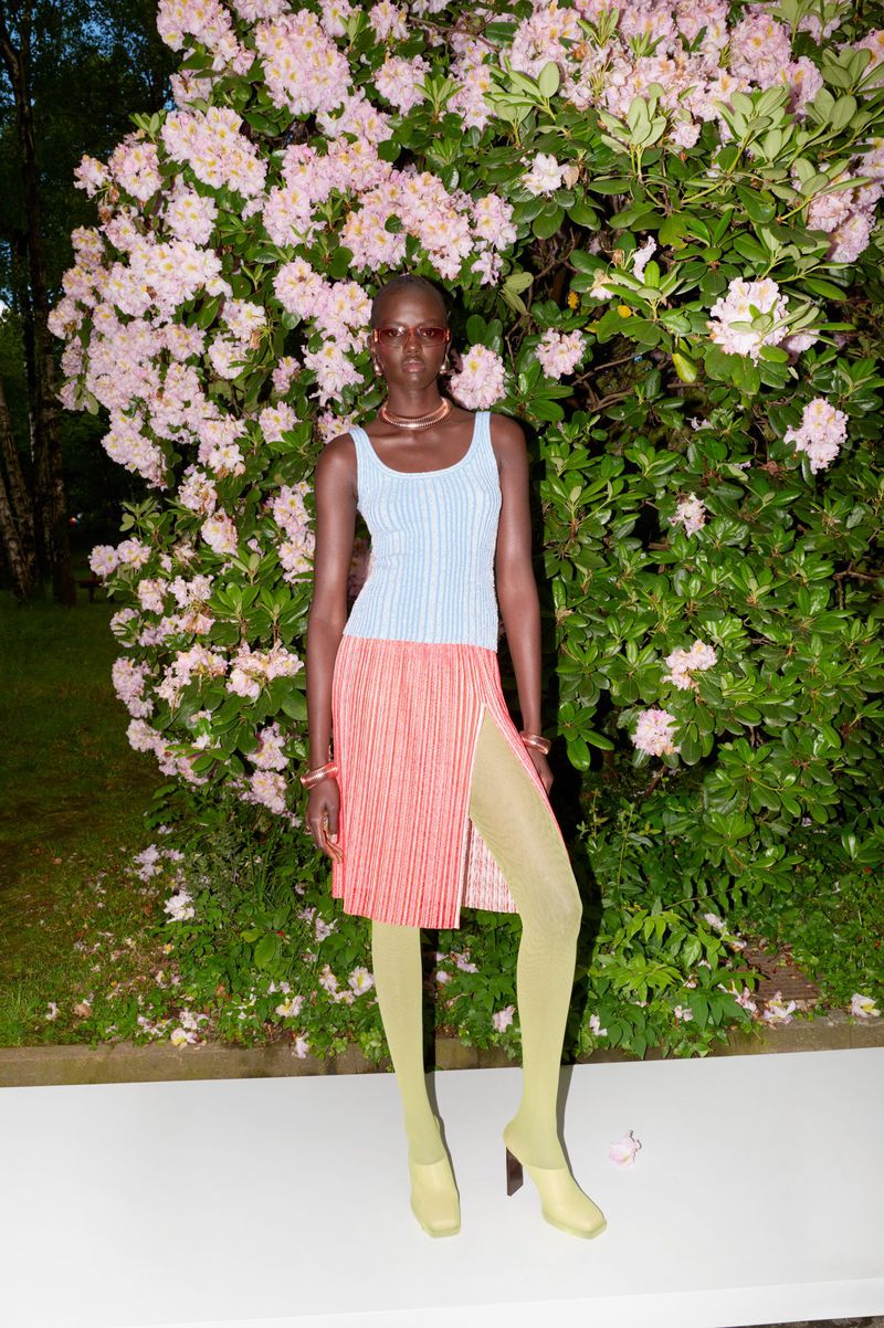 Woman in blue top and coral skirt poses in front of flowering bush. Full shot in natural lighting. Fashion editorial photography.