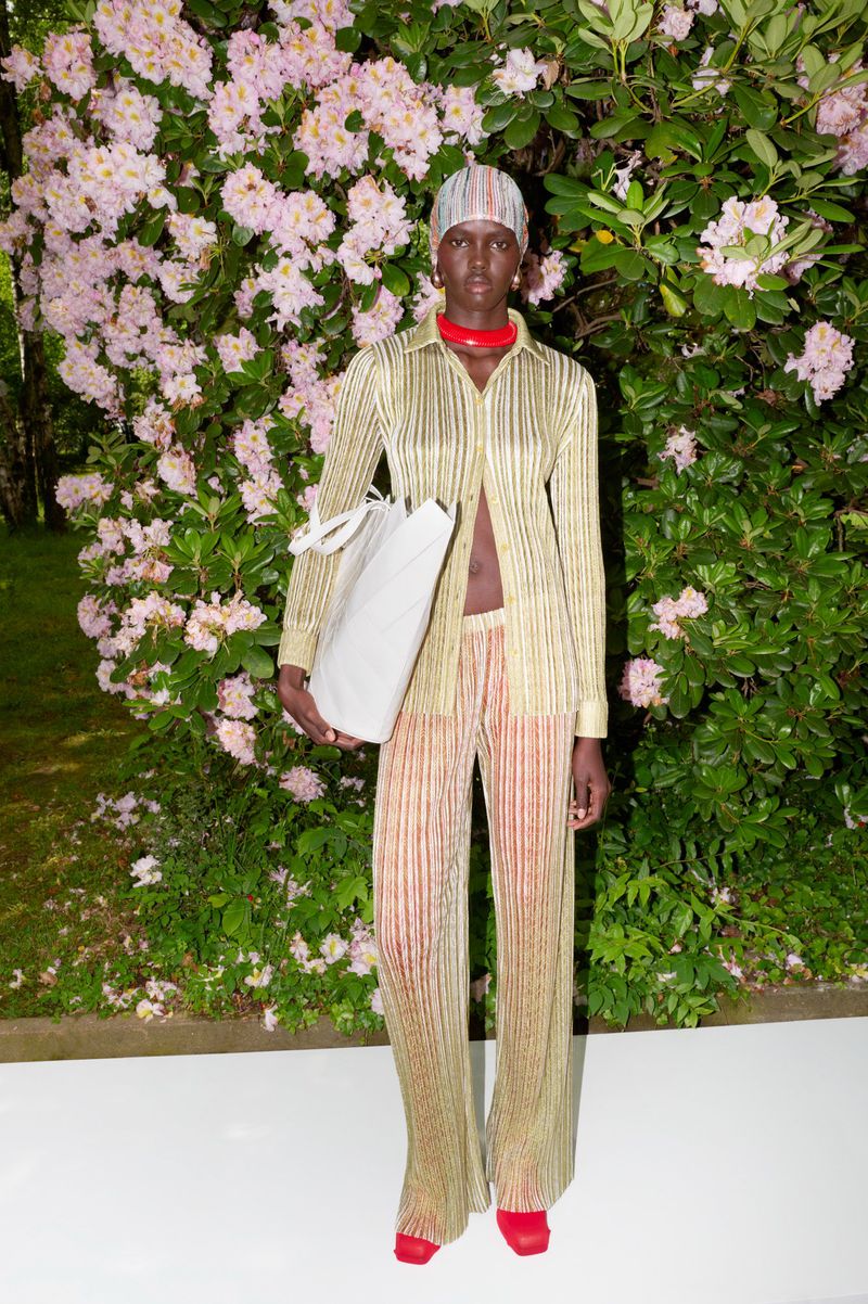 Elegant woman in a gold striped shirt and pants stands outside with a white tote bag, posing in front of flowering bushes.