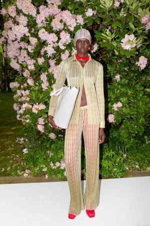 Elegant woman in a gold striped shirt and pants stands outside with a white tote bag, posing in front of flowering bushes.